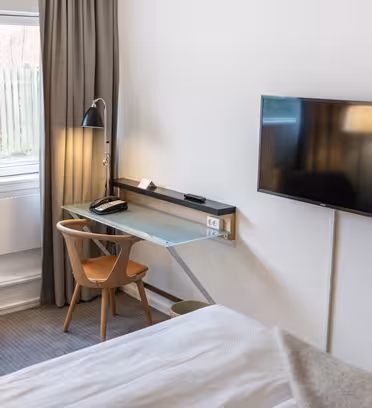 Et moderne hotelværelse med en stor seng, et skrivebord med glasplade og en træstol. Et fladskærms-tv er monteret på væggen, og en gulvlampe er ved siden af skrivebordet. Rummet har vindue med grå gardiner og udsigt til et plankeværk udenfor.