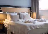 Et moderne hotelværelse har en pænt lavet dobbeltseng med hvidt sengetøj, beige puder og et lysegråt tæppe. To badekåber er lagt ud på sengen. Rummet er blødt oplyst af to væghængte lamper, med gardiner, der indrammer et vindue.
