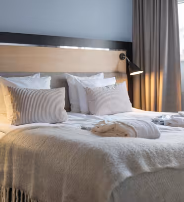 Et moderne hotelværelse har en pænt lavet dobbeltseng med hvidt sengetøj, beige puder og et lysegråt tæppe. To badekåber er lagt ud på sengen. Rummet er blødt oplyst af to væghængte lamper, med gardiner, der indrammer et vindue.