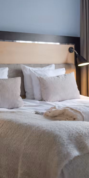 Et moderne hotelværelse har en pænt lavet dobbeltseng med hvidt sengetøj, beige puder og et lysegråt tæppe. To badekåber er lagt ud på sengen. Rummet er blødt oplyst af to væghængte lamper, med gardiner, der indrammer et vindue.