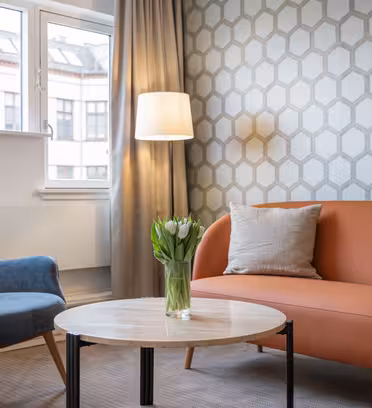 En moderne stue med et sekskantet mønstret tapet, med en orange sofa og en blå lænestol. Et rundt sofabord i træ rummer en vase med tulipaner, og en gulvlampe står ved siden af sofaen, nær et vindue med gardiner.