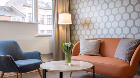 En moderne stue med et sekskantet mønstret tapet, med en orange sofa og en blå lænestol. Et rundt sofabord i træ rummer en vase med tulipaner, og en gulvlampe står ved siden af sofaen, nær et vindue med gardiner.