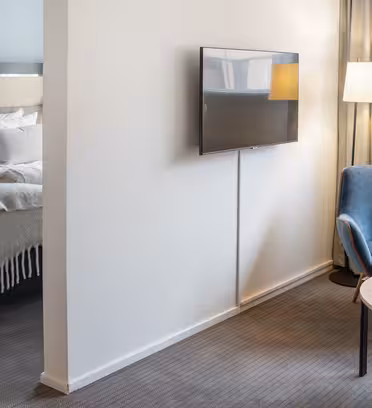 Moderne hotelværelse med en dobbeltseng i baggrunden, et fladskærms-tv på en hvid væg og en hyggelig blå stol nær et rundt træbord med en vase med tulipaner. Blød belysning fra en gulvlampe og naturligt lys fra vinduet forstærker stemningen.