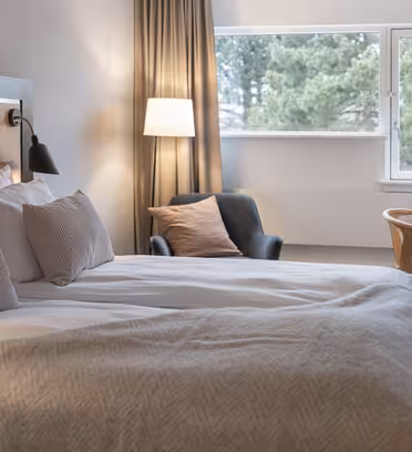 Et hyggeligt hotelværelse med en stor seng, bløde puder og et beige tæppe. Der er en sort lænestol, et skrivebord med en stol, en lampe og et vindue, der viser grønt udenfor. Neutrale toner skaber en rolig, indbydende atmosfære.
