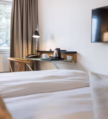 Hotelværelse med en pæn opredt seng, et væghængt tv og et skrivebord ved vinduet. Skrivebordet har en moderne lampe, en stol og en kedel med kopper. Gardiner dækker en del af vinduet, og træer kan ses udenfor.