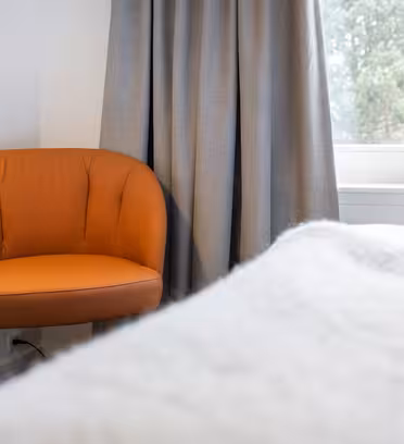 En orange stol sidder i et hjørne ved et vindue med grå gardiner. Et hvidt tæppe er synligt i forgrunden, hvilket antyder et hyggeligt rum. Træer kan ses uden for vinduet.