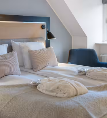 Hyggeligt hotelværelse med en pænt lavet dobbeltseng med hvidt sengetøj og beige accentpuder. To morgenkåber er placeret på sengen. En blå lænestol er tæt på et vindue med byudsigt, og en vægmonteret lampe giver lys.