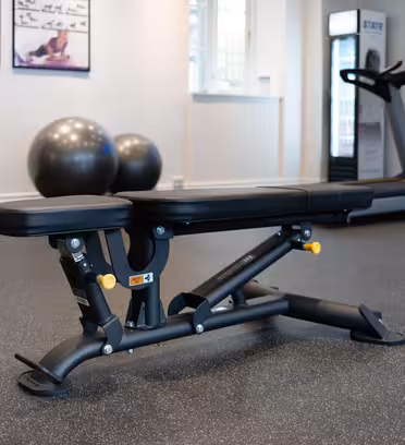 Et fitnesscenter-interiør, der viser en justerbar sort træningsbænk på et gråt plettet gulv. I baggrunden ses to store træningsbolde, et løbebånd, en plakat med øvelser og en del af et vægtstativ.
