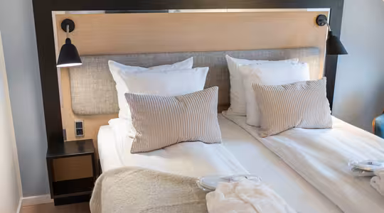 Et pænt indrettet hotelværelse har to senge med hvidt sengetøj og beige accentpuder. Sengegavlen har påsat sorte læselamper, og et natbord rummer faciliteter. En foldet morgenkåbe er placeret ved foden af den ene seng.