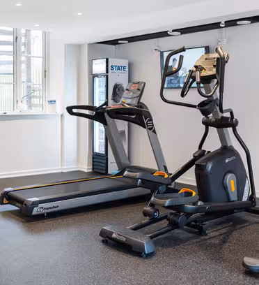 Et moderne fitnessrum med et løbebånd, en elliptisk, stationær cykel og træningsbold. Et fladskærms-tv er monteret på væggen, og en salgsautomat er i nærheden. Rummet har et stort vindue og et mørkt gummigulv.