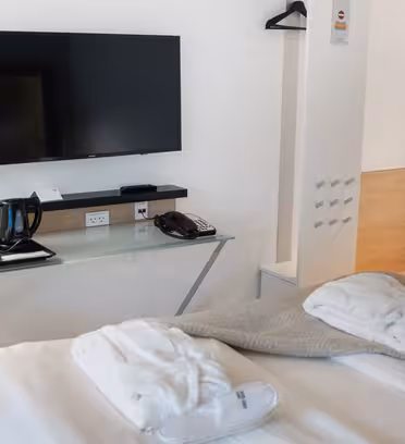 Et moderne hotelværelse har et vægmonteret tv, et skrivebord med en lampe, tefaciliteter og en væghængt telefon. To badekåber er pænt placeret på sengen. En vævet kurv sidder under skrivebordet, og en bøjle er monteret på væggen.