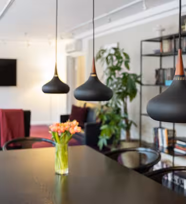 En moderne stue med tre sorte pendellamper hængende over et træbord. En vase med orange tulipaner står på bordet. I baggrunden er der en bogreol, et fladskærms-tv, stole med røde plader og en høj plante.