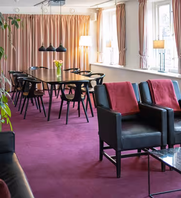 En moderne stue med et langt spisebord, sorte stole og hængende lys. To sorte lænestole med røde hynder vender mod et marmor sofabord. En sort sofa er til venstre. Pink tæppe og gardiner, store vinduer og en høj plante fuldender scenen.