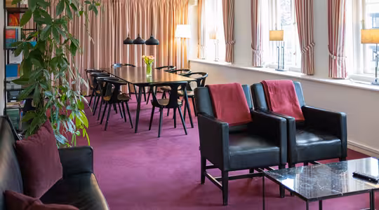 En moderne stue med et langt spisebord, sorte stole og hængende lys. To sorte lænestole med røde hynder vender mod et marmor sofabord. En sort sofa er til venstre. Pink tæppe og gardiner, store vinduer og en høj plante fuldender scenen.