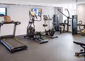 Et moderne fitnessrum med forskellige træningsredskaber, herunder et løbebånd, elliptiske maskiner, stationær cykel og styrketræningsmaskiner. Et TV er monteret på den hvide væg. Gulvet er belagt med mørk plettet gummimåtte.
