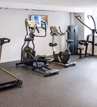 Et moderne fitnessrum med forskellige træningsredskaber, herunder et løbebånd, elliptiske maskiner, stationær cykel og styrketræningsmaskiner. Et TV er monteret på den hvide væg. Gulvet er belagt med mørk plettet gummimåtte.
