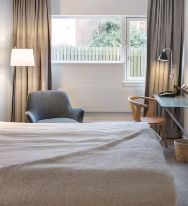 Et moderne hotelværelse har en pænt redt seng med hvidt sengetøj, en hyggelig grå lænestol, et fladskærms-tv monteret på væggen, en træstol med et glasbord under tv'et og et stort vindue med grå gardiner, der slipper naturligt lys ind.