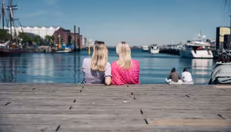 To personer med langt blondt hår sidder side om side på en trædok og vender ud mod en rolig havnefront med både og bygninger i det fjerne. Personen til venstre bærer lys lilla; personen til højre bærer lyserødt. Klar blå himmel over hovedet.