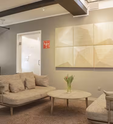 Et moderne venteværelse med to beige sofaer over for hinanden, et rundt sofabord i træ med en vase med blomster, tekstureret vægkunst, et lysebrunt tæppe og et gardin. Rummet har et minimalistisk design med neutrale toner.