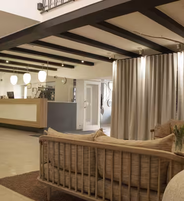 En moderne lobby med beige sofaer, en reception i træ og hængende runde lamper. En vindeltrappe er synlig til venstre, og der er en stor vase med kviste i forgrunden. Neutrale farver og gardiner bidrager til den hyggelige stemning.