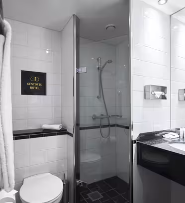 Hotelbadeværelse med hvide flisevægge, walk-in glasbruser og vægmonteret brusehoved. En sort bordvask med toiletartikler og et spejl er synlig. Rullede håndklæder er stablet i nærheden af et toilet. På et skilt står der "Gentofte Hotel.
