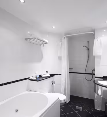 Et moderne badeværelse med et hvidt badekar, en hjørnebruser med et gardin, et toilet og en håndvask med spejl og håndvask. Rummet er flisebelagt i hvidt med sort kant og har indbyggede loftslamper.