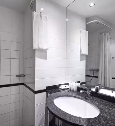 Moderne badeværelse med hvide flisevægge med bruseniche med gardin og sort granitbordplade med oval vask. Håndklæder hænger på væggen, og toiletartikler er pænt arrangeret ved håndvasken. Et stort spejl afspejler rummet.