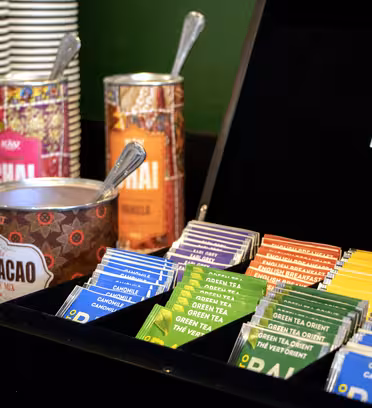 Assorterede teposer er organiseret i en sort boks med forskellige smagsvarianter som kamille og grøn te. Bag den er der farverige dåser mærket "Chai" og "Cacao", med stablede hvide kopper og en ske i forgrunden.