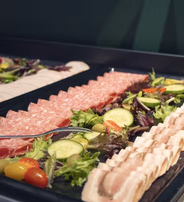 Et fad med diverse delikatesser, inklusive salami og skiver kalkun, arrangeret i rækker på en bakke. Frisk grønt, agurk i skiver, cherrytomater og oliven lægges mellem rækkerne. En serveringsske hviler på bakken.