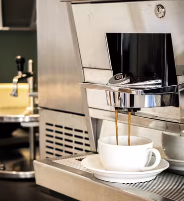 Et nærbillede af en kaffemaskine, der serverer espresso i en hvid kop og underkop. Maskinen er metallisk og moderne, med en poleret finish. I baggrunden er der utydelige køkkenting, blandt andet en skinnende kaffekande.