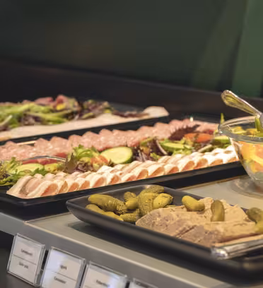 Et buffetbord med bakker med diverse skiveskåret kød og ost, pyntet med salat, cherrytomater og agurker. Der er også en skål med blandet frugt og et fad med pickles. Skilte med etiketter er synlige under bakkerne.