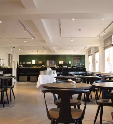 Rummeligt, moderne cafeteria med træborde og stole. Glasdøre til venstre og store vinduer til højre slipper naturligt lys ind. En servicedisk med kaffemaskiner og selvbetjeningsartikler er synlig i baggrunden.