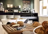 Et hyggeligt morgenmadssetup med skiveskåret brød og kager på træplader. En kniv hviler på et skærebræt. I baggrunden rummer hylder tallerkner og kopper, med et tavleskilt, der byder gæster velkommen. Lyset strømmer ind gennem et nærliggende vindue.