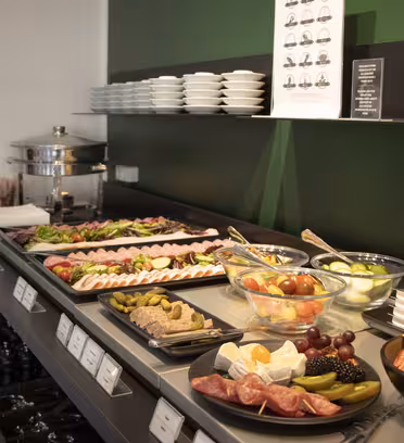 Et buffetbord med en række forskellige fødevarer, herunder kød i skiver, ost, frugt, oliven og salater i skåle. Tallerkener og småretter er stablet på en hylde ovenover. En sort tæller med en grøn væg i baggrunden fuldender opsætningen.