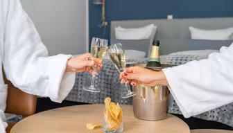 To personer i hvide rober skåler med champagneglas på et stilfuldt hotelværelse. En flaske champagne hviler i en isspand på bordet sammen med et glas fyldt med kartoffelchips. Plys sengetøj er synligt i baggrunden.